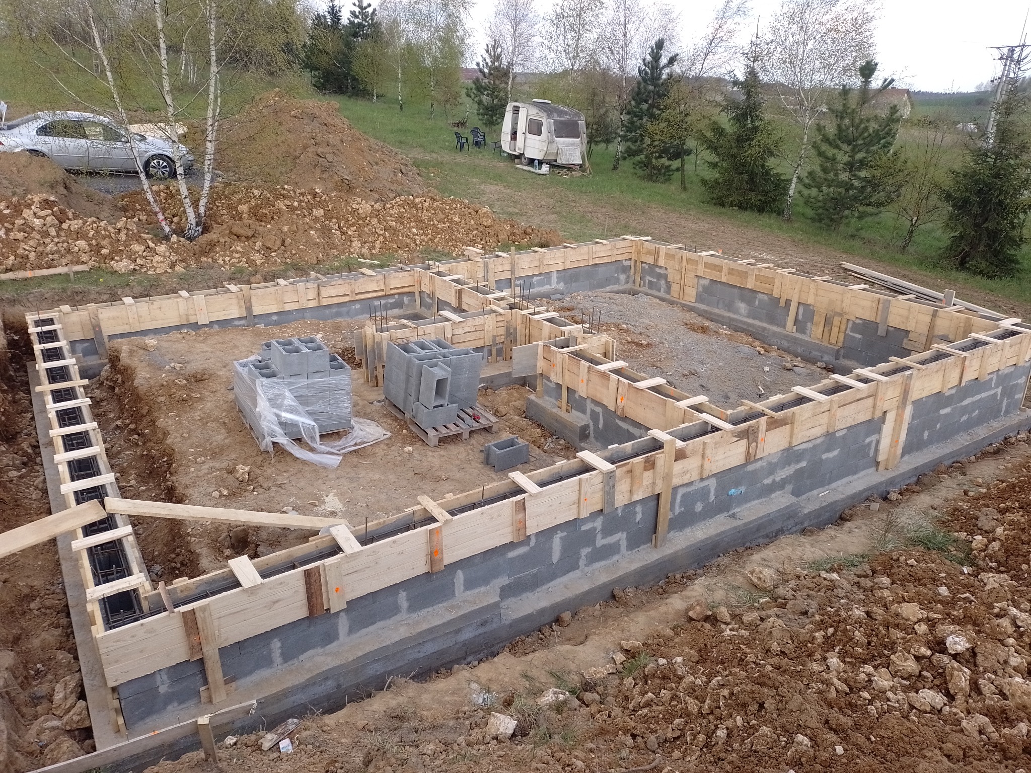 Fundamenty domu jednorodzinnego w trakcie budowy, szalunek z desek i bloczków betonowych, w tle przyczepa kempingowa i samochód osobowy.