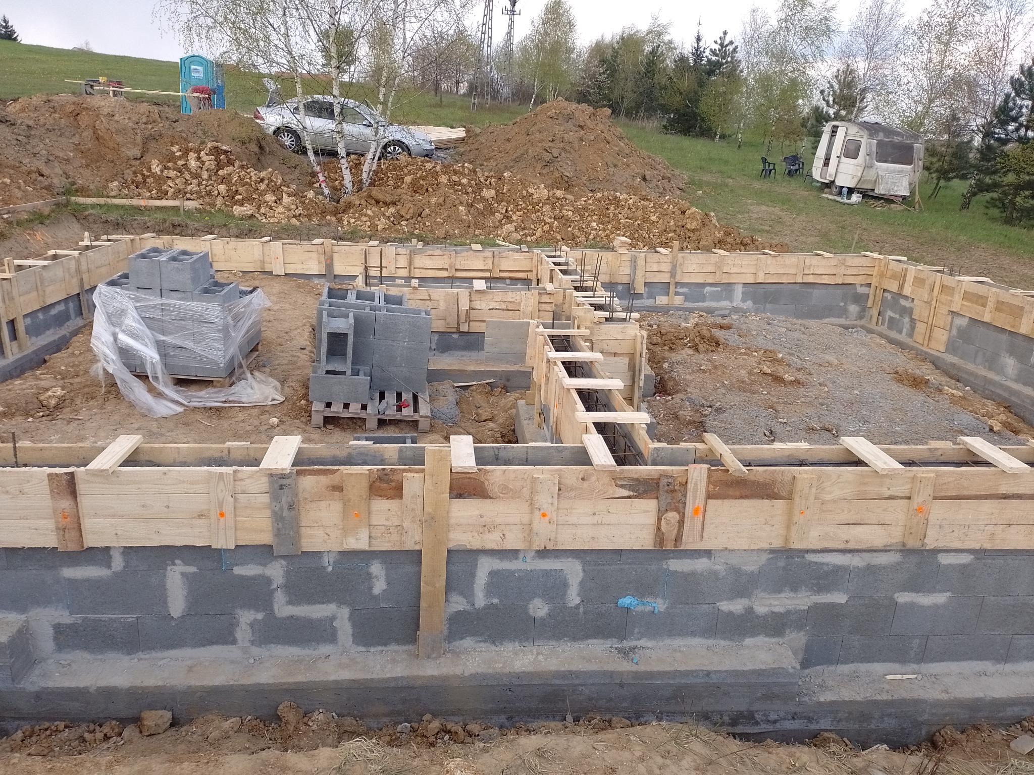 Wykopane fundamenty z szalunkiem drewnianym i częściowo wzniesionymi ścianami z betonowych bloczków, widoczny stos bloczków na palecie oraz prowizoryczna toaleta i przyczepa kempingowa w tle.