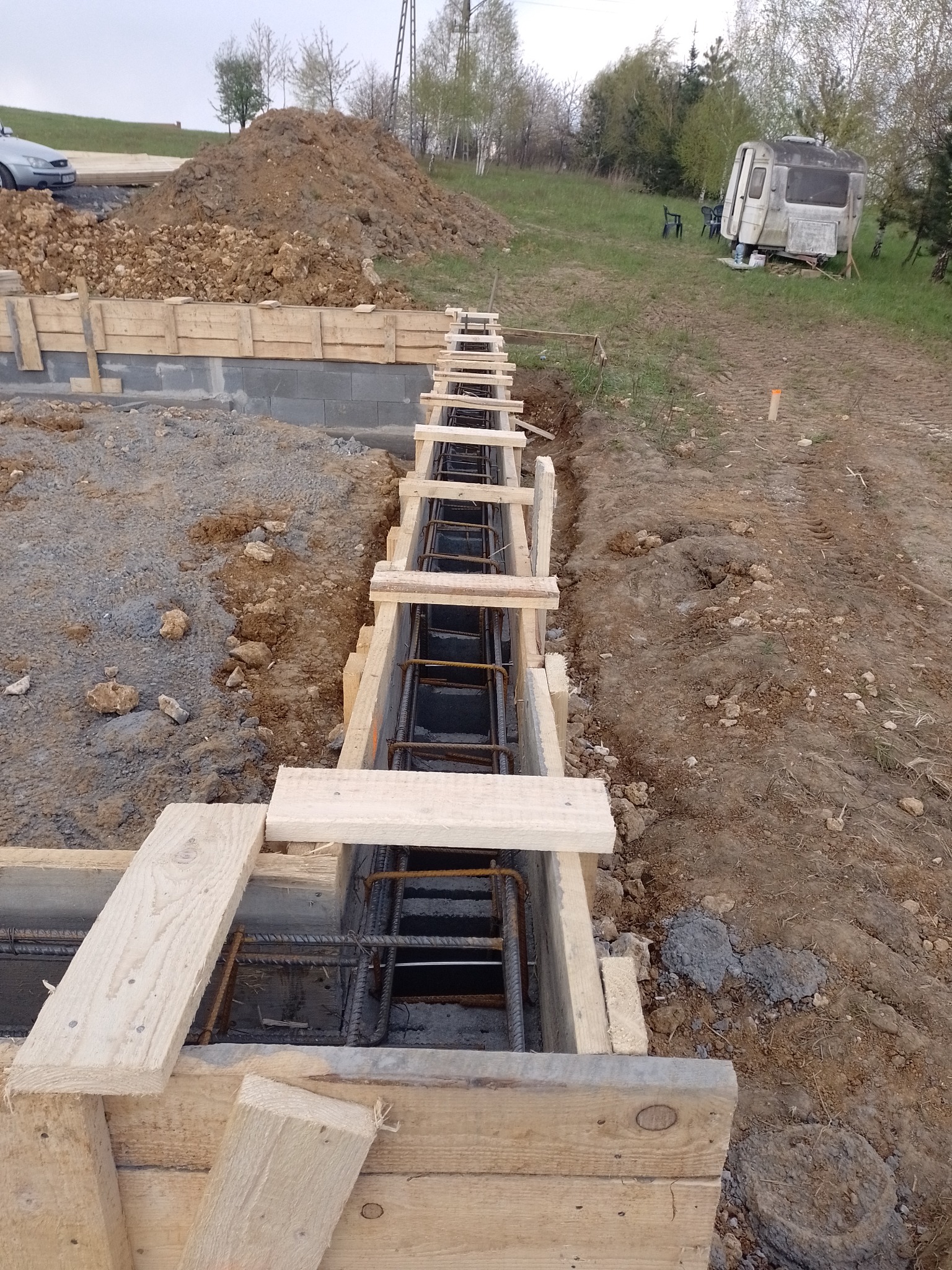 Zbrojenie w szalunku drewnianym przygotowane do zalania betonem pod fundament domu jednorodzinnego, w tle ziemia, przyczepa kempingowa i zaparkowany samochód.