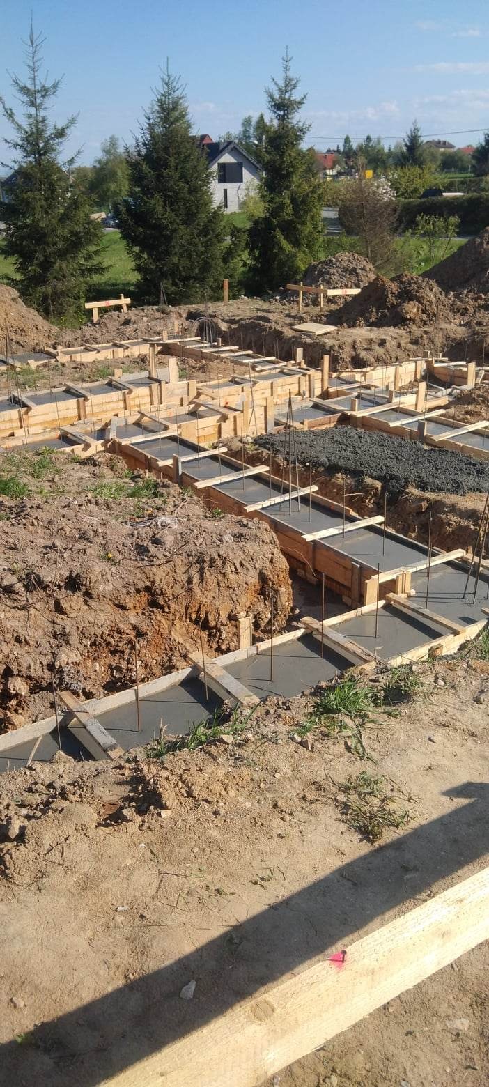 Przygotowane szalunki i zalane betonem ławy fundamentowe pod budowę domu jednorodzinnego, widoczne wystające pręty zbrojeniowe oraz otaczający teren budowy.