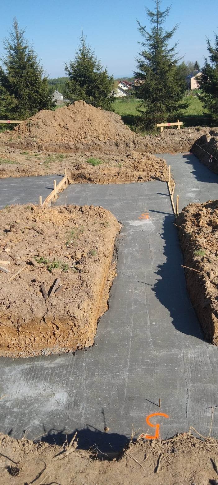Wylane fundamenty pod budynek, widoczne drewniane szalunki i wykopana ziemia, w tle drzewa iglaste i zabudowania.