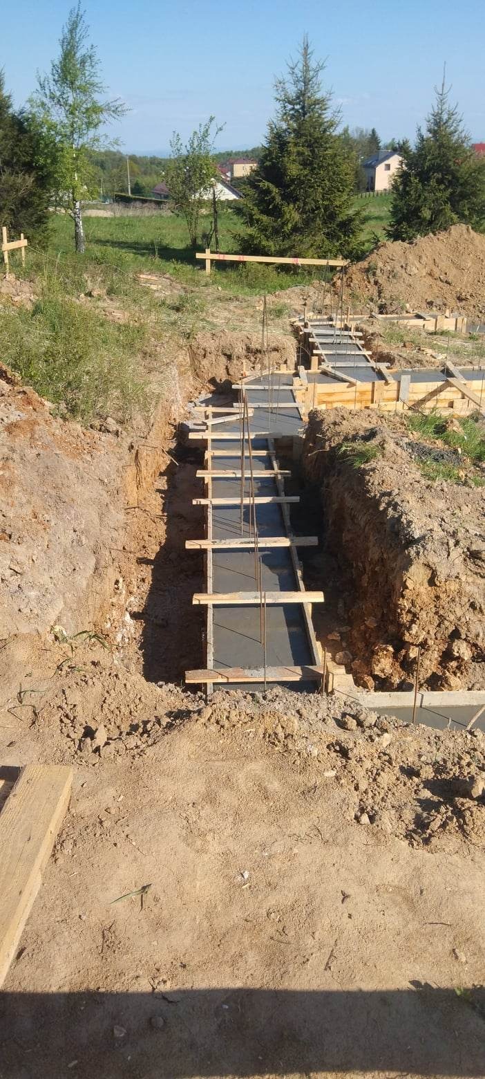 Zalewanie betonem szalunków pod fundamenty domu jednorodzinnego, widoczne zbrojenie pionowe i drewniane rozpory.