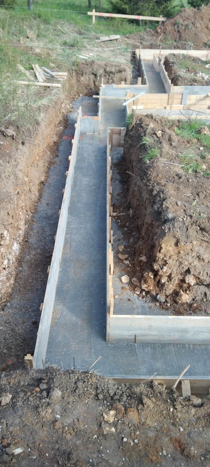 Zalane betonem szalunki pod fundamenty domu jednorodzinnego, widoczne zbrojenia i wykopane rowy w ziemi.