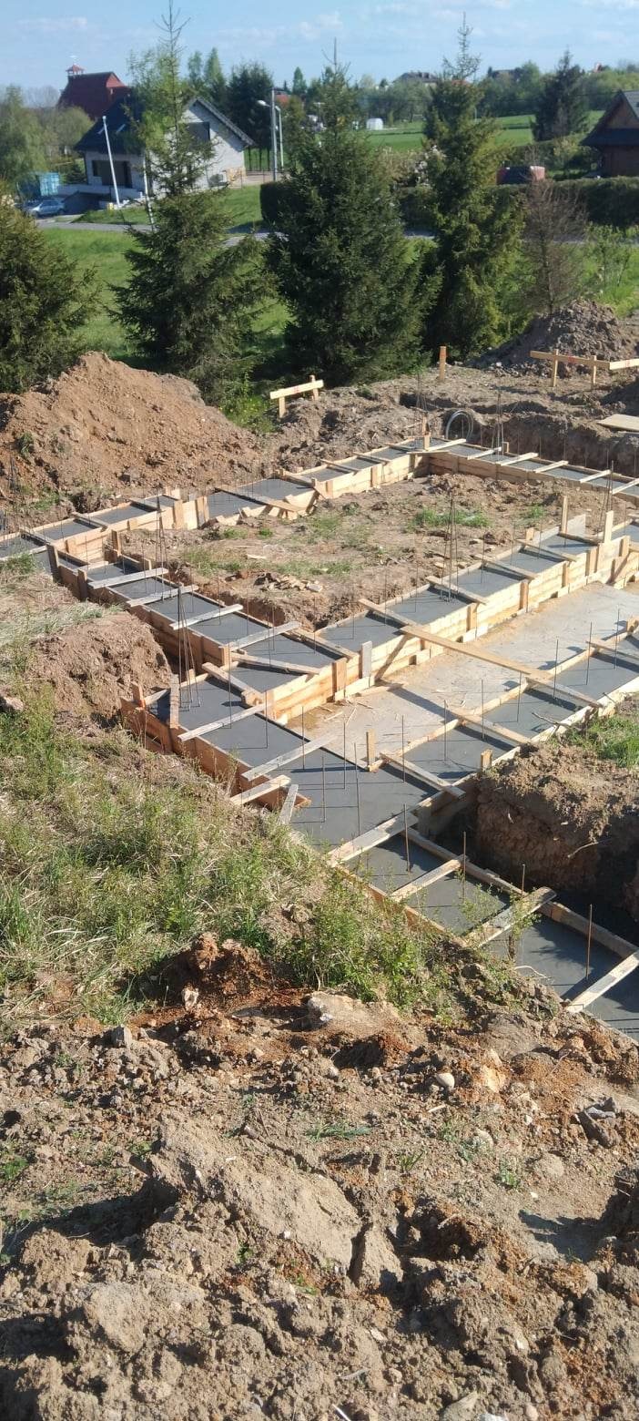 Wykopane fundamenty z szalunkiem drewnianym, częściowo zalane betonem z widocznym zbrojeniem, w tle zielona trawa i dom w budowie.