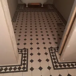 Ukończona łazienka z wolnostojącą wanną na tle nowej, czarno-białej podłogi w karo z ozdobnym obramowaniem, widok z góry na fragment drzwi.