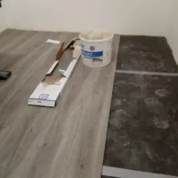 Układanie paneli podłogowych z widoczną otwartą paczką paneli, wiaderkiem po farbie i fragmentem starej podłogi z śladami stóp, na etapie prac wykończeniowych.