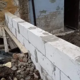 Wznoszenie ściany z bloczków betonowych, widoczne świeże spoiny cementowe, gruz u podstawy i fragment starej, kamiennej ściany w tle.