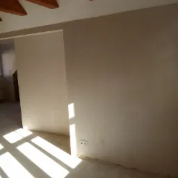 Pokój w trakcie prac przygotowawczych do wykończenia: gładkie, jasne ściany, surowy beton na podłodze, widoczne drewniane belki stropowe i cień wpadający przez okno.