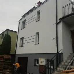 Dwupiętrowy dom po malowaniu elewacji na biało-szaro z numerem 13, widoczne rusztowania i pracownik w żółtej kurtce przy garażu.