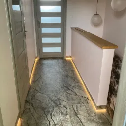 Korytarz z marmurową posadzką, oświetleniem LED przy podłodze, różowymi ścianami i dwiema drewnianymi ościeżnicami drzwi.