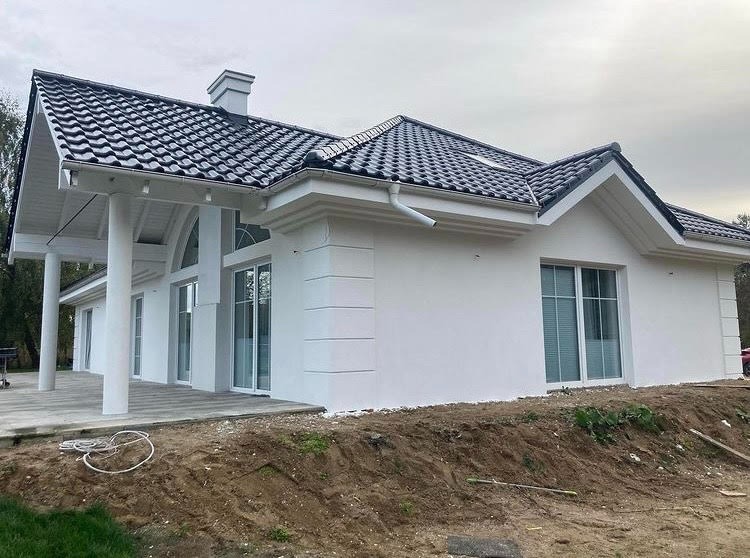 Nowoczesny dom jednorodzinny z białą elewacją, czarną dachówką i kolumnowym gankiem, widoczny na tle pochmurnego nieba, z niezagospodarowanym terenem wokół.