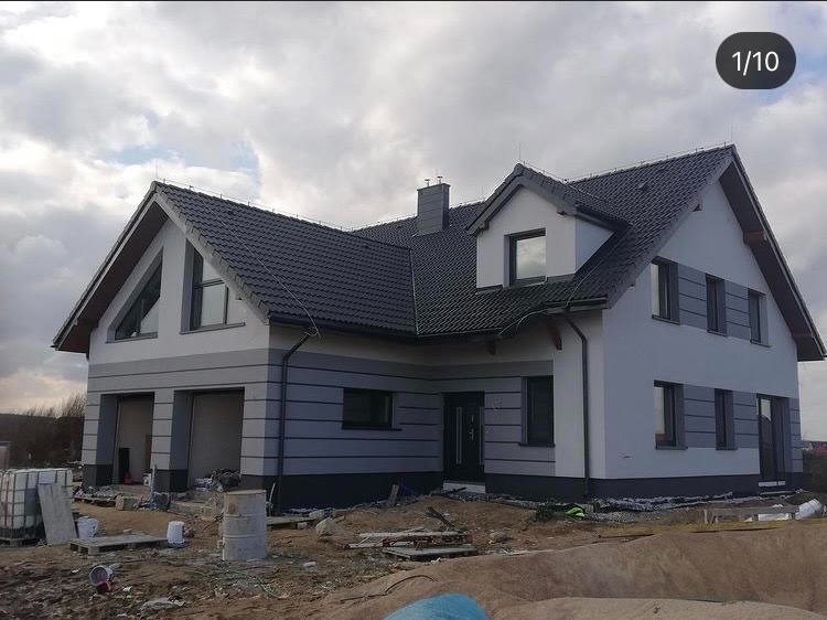 Dwukondygnacyjny dom z szarą elewacją, ciemnoszarą stolarką okienną i dachem z ciemnej dachówki, teren wokół domu w trakcie prac wykończeniowych.