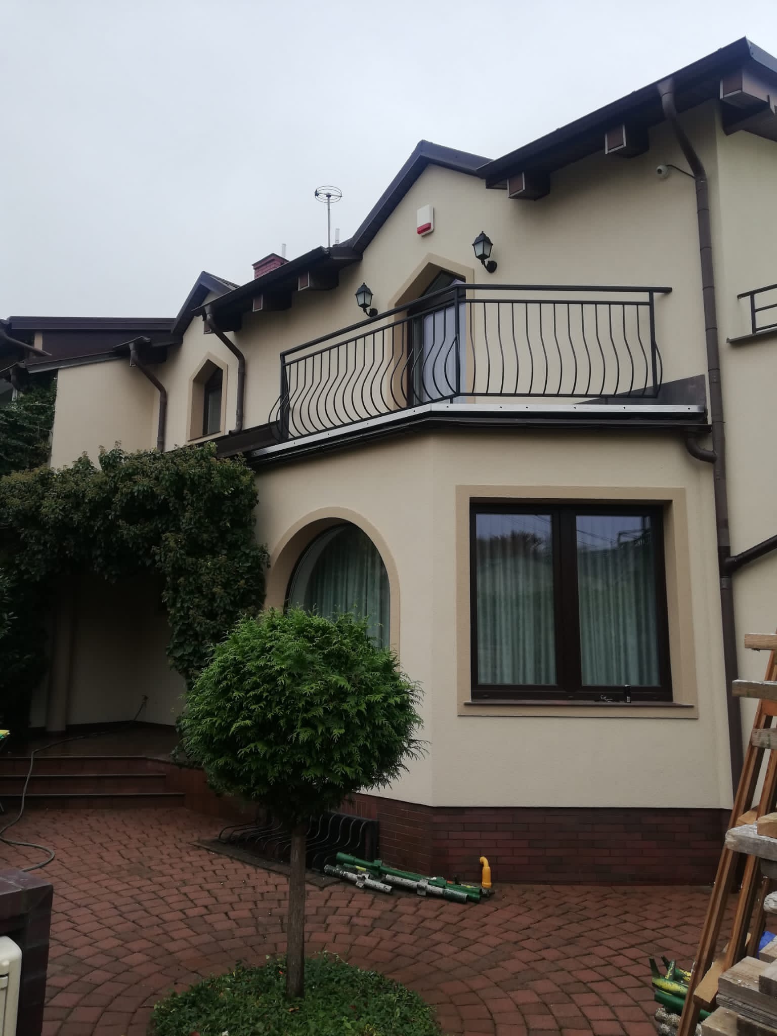 Jasna elewacja domu jednorodzinnego z balkonem o metalowej balustradzie, widoczne elementy wykończeniowe w kolorze brązowym, małe drzewko iglaste i fragment drabiny.