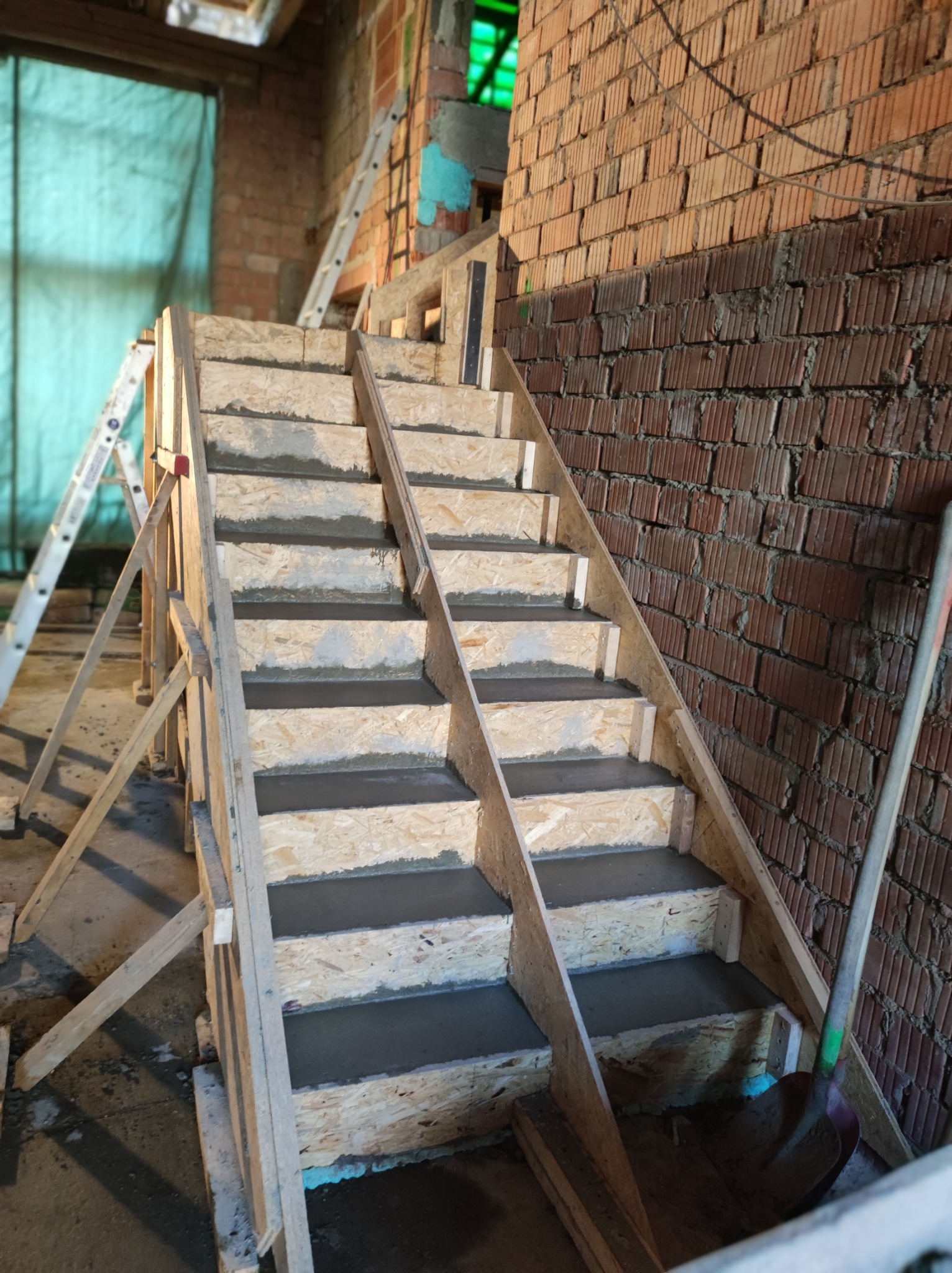 Surowe, drewniane szalunki schodów wypełnione świeżym betonem w surowym wnętrzu budynku z ceglaną ścianą i drabiną w tle.