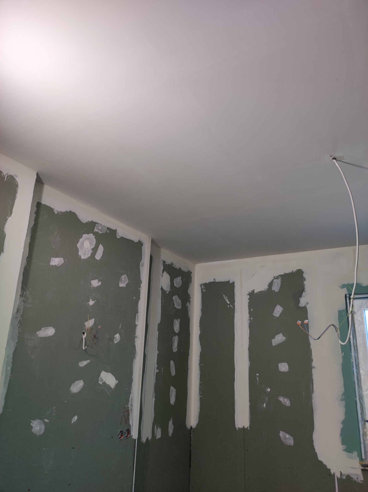 Narożnik pokoju w trakcie szpachlowania płyt kartonowo-gipsowych, widoczne łączenia płyt i miejsca po wkrętach pokryte gładzią, sufit pomalowany na biało, widoczne przewody elektryczne.