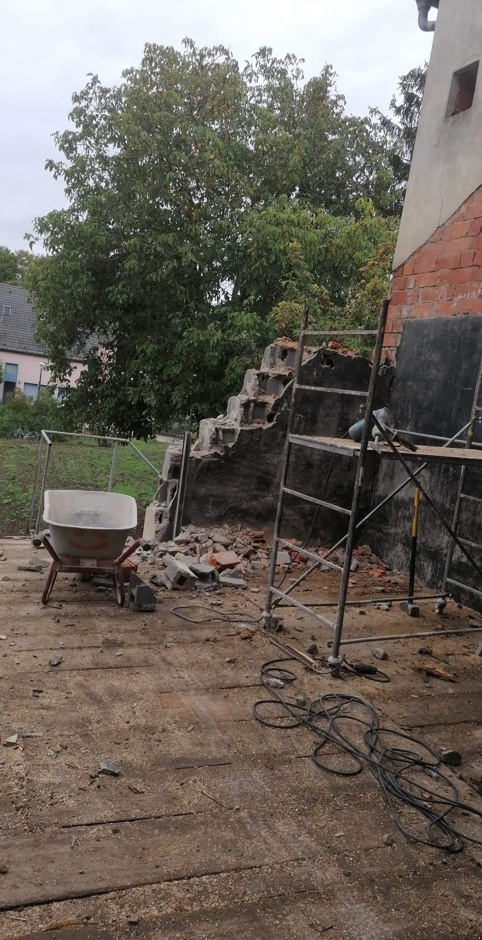 Fragment zdemontowanej ściany z cegły i betonu, gruz, taczka z przodu, rusztowanie z boku, widok na drzewo i okoliczne budynki w tle.