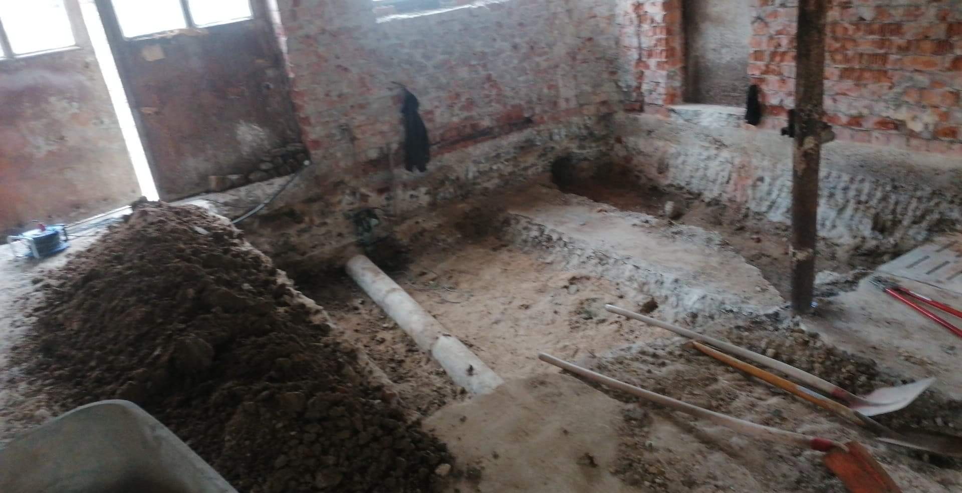 Wykop wewnątrz budynku z odsłoniętą cegłą, widoczne rury, łopaty i sterta ziemi, przygotowanie do prac instalacyjnych.
