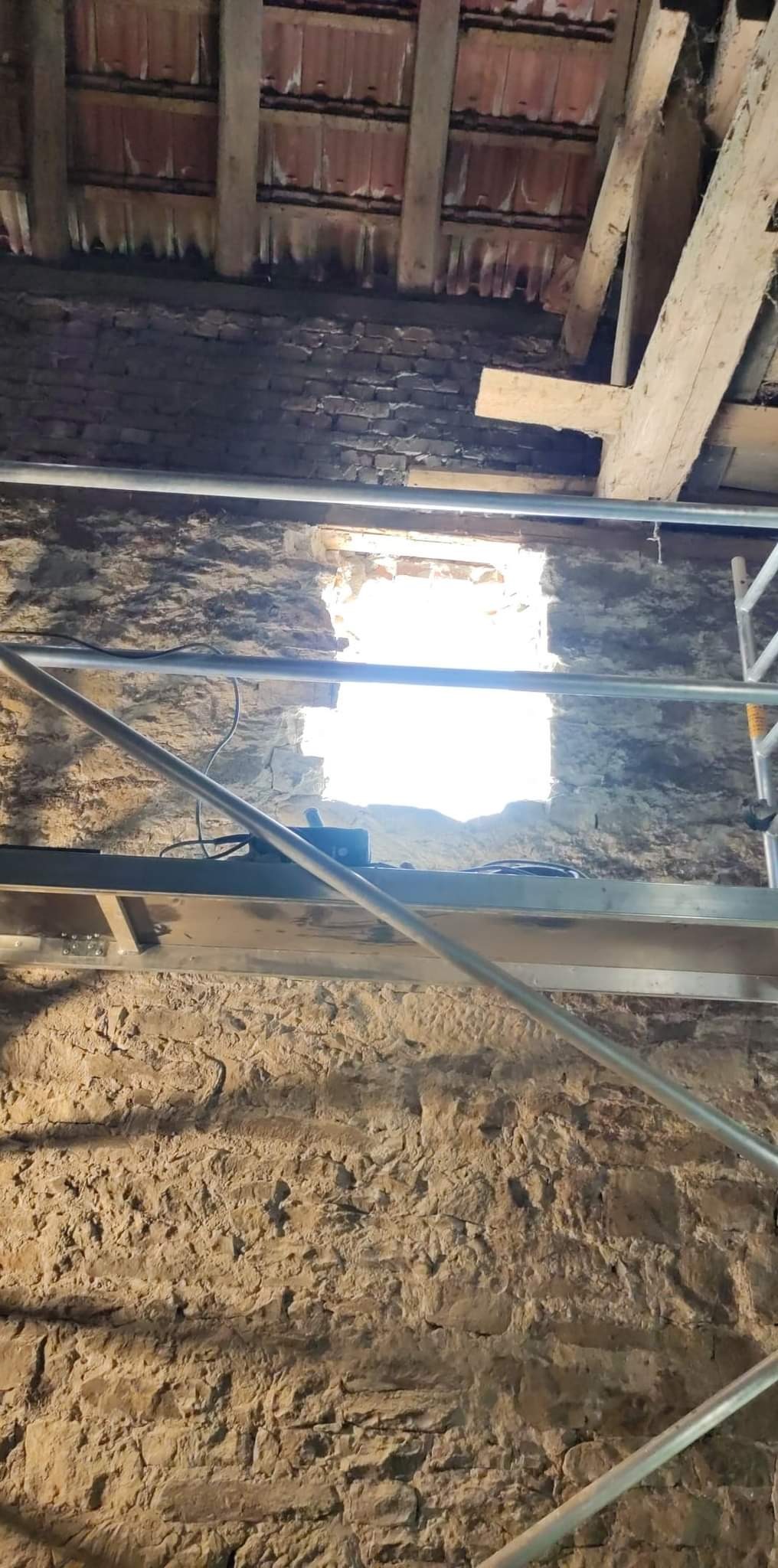 Surowa kamienna ściana wewnętrzna z widocznym otworem okiennym w trakcie prac, z fragmentem rusztowania na pierwszym planie i drewnianą konstrukcją dachu w tle.