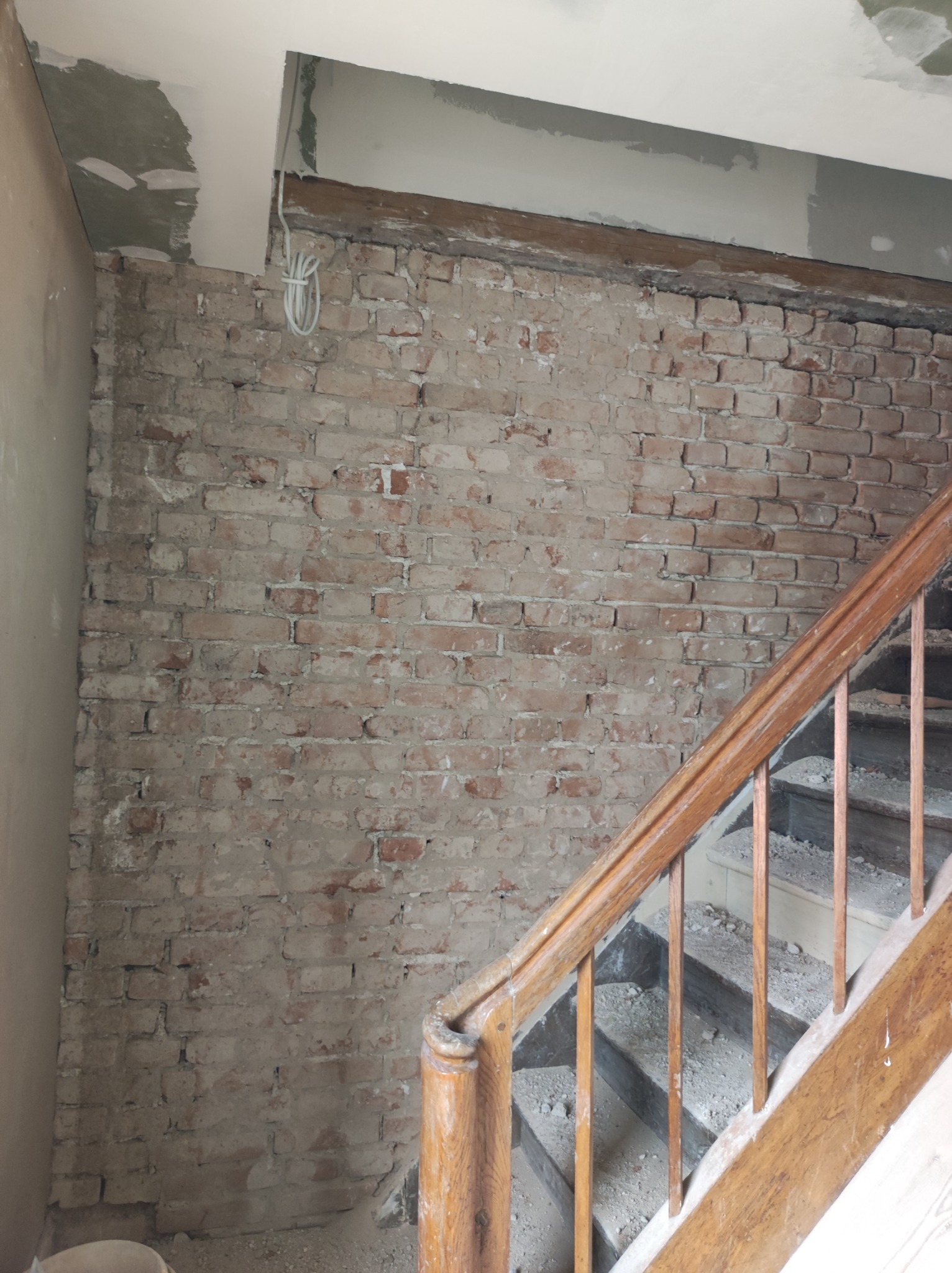 Ceglana ściana w trakcie remontu, widoczne fragmenty starego muru, drewniane schody z zabrudzeniami, oraz fragmenty instalacji elektrycznej pod sufitem.