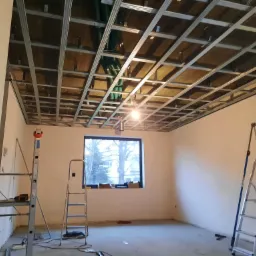 Montaż metalowej konstrukcji sufitu podwieszanego z widocznym ociepleniem i przewodami wentylacyjnymi, drabiny w pustym pomieszczeniu z oknem.