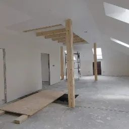 Poddasze w trakcie prac wykończeniowych, widoczne drewniane podpory, płyta OSB na podłodze, ściany po wstępnym wyr&oacute;wnaniu, okna dachowe wpuszczające światło.