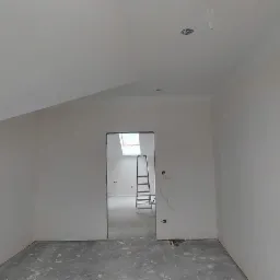 Poddasze po remoncie: białe ściany, gładzie, widoczna drabina w przejściu do kolejnego pomieszczenia, niebieski przewód wentylacyjny na ścianie.