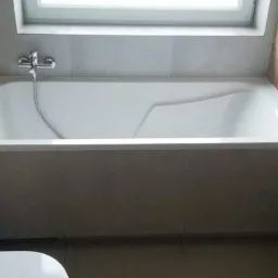 Nowoczesna biała wanna zabudowana w szarej obudowie, z chromowaną baterią, zamontowana w łazience wykończonej płytkami imitującymi drewno i szarym gresem.