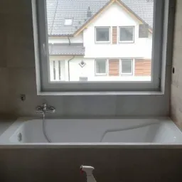 Nowoczesna łazienka z wanną obudowaną płytkami imitującymi drewno, widok na dom przez okno, chromowana bateria wannowa.