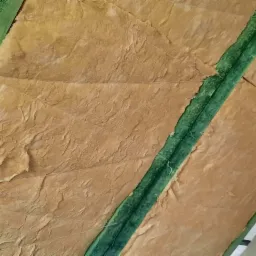 Poddasze ocieplone wełną mineralną, widoczne zielone belki konstrukcyjne i białe sznurki mocujące izolację, fragment ściany z gładzią.