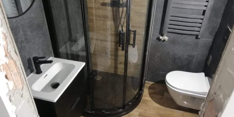 Nowoczesna łazienka z czarnymi akcentami: kabina prysznicowa z ciemną ramą, umywalka z czarną armaturą, wisząca toaleta i szary grzejnik drabinkowy na tle ciemnych płytek, widoczne ślady po remoncie.