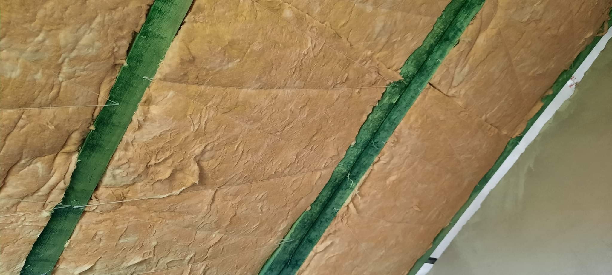 Poddasze ocieplone wełną mineralną, widoczne zielone belki konstrukcyjne i białe sznurki mocujące izolację, fragment ściany z gładzią.