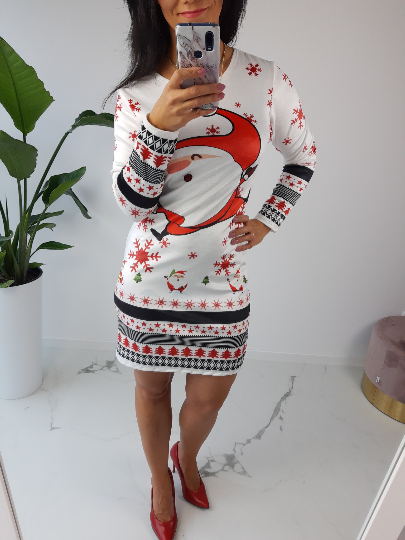 Kobieta robi selfie w świątecznej tunice z motywem Mikołaja, stojąc w jasnym wnętrzu w Woli Mrokowskiej. Widoczne czerwone szpilki i doniczka z rośliną.