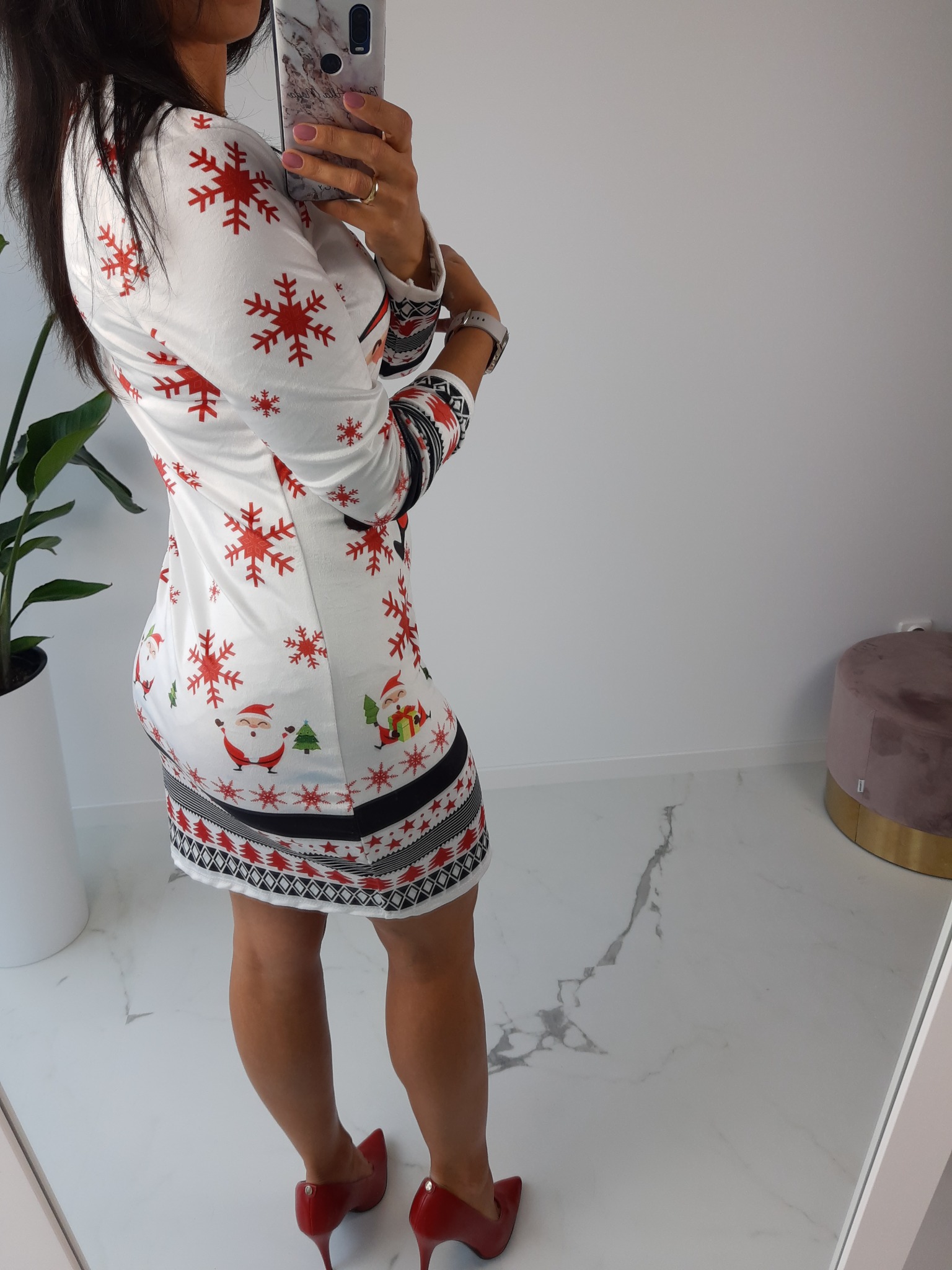 Kobieta robi selfie w świątecznej sukience z motywem śnieżynek i Mikołaja, pozując w czerwonych szpilkach na marmurowej podłodze.