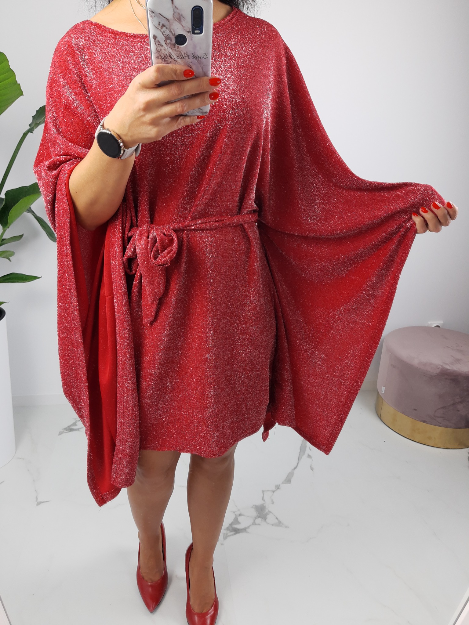 Kobieta prezentuje czerwoną, błyszczącą sukienkę z wiązaniem w pasie, robiąc sobie zdjęcie telefonem z marmurkowym etui, widoczne czerwone szpilki i minimalistyczny zegarek.