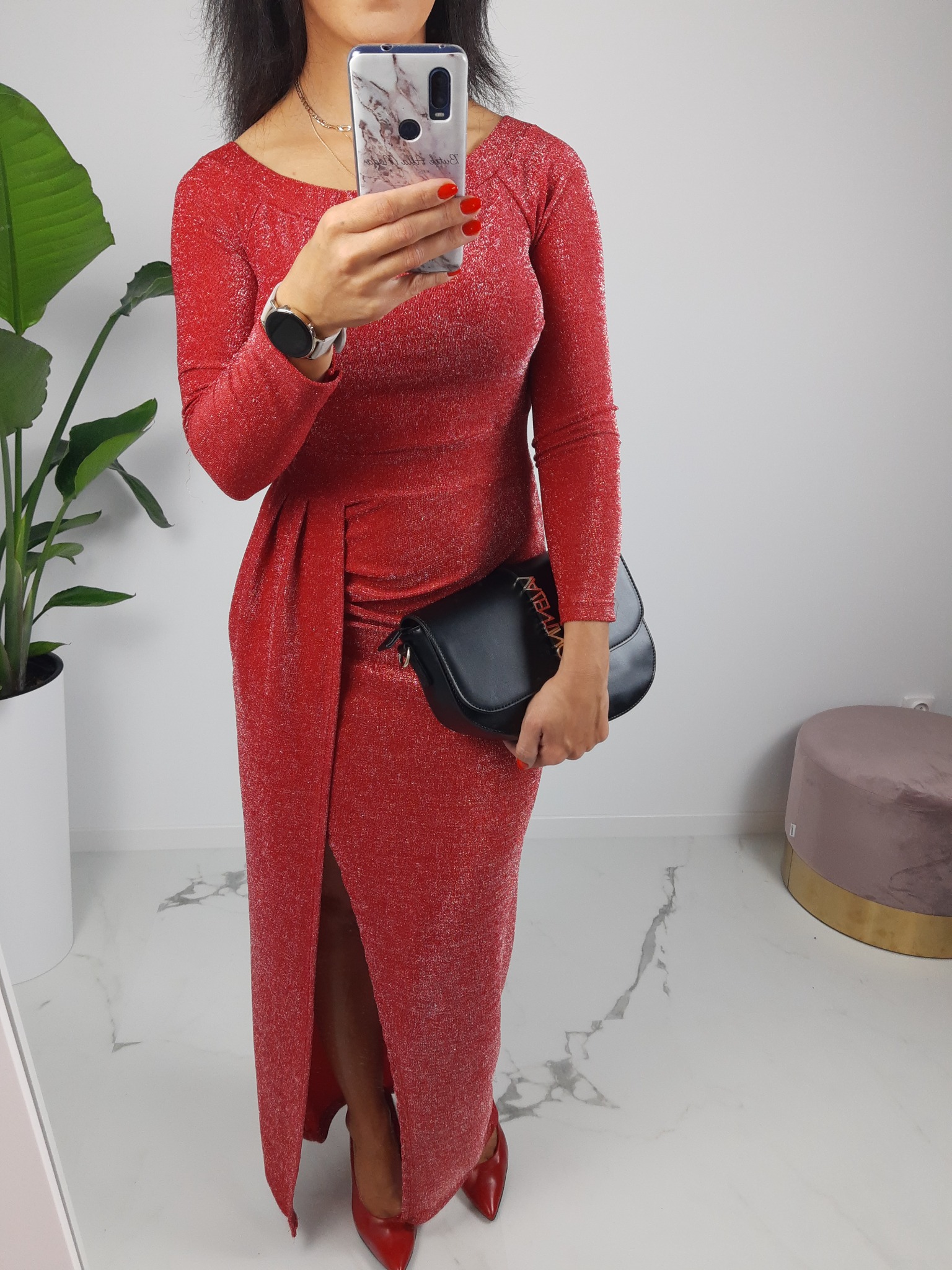 Kobieta robi selfie w czerwonej, brokatowej sukience z rozcięciem, trzymając czarną torebkę z ozdobnym napisem; widoczne czerwone szpilki, zegarek na ręce i marmurkowe etui na telefon.