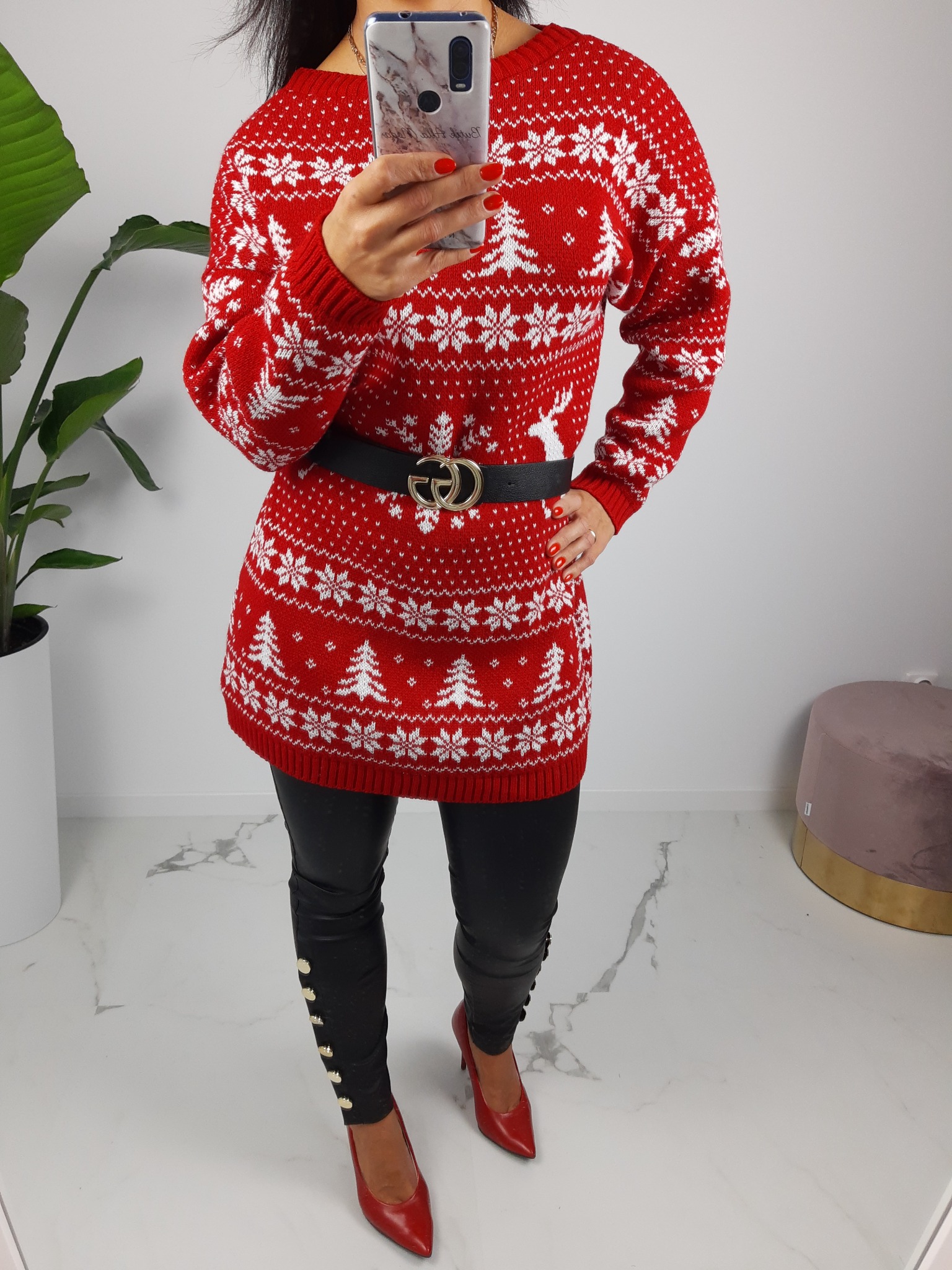 Czerwona dzianinowa tunika z białymi motywami świątecznymi (renifery, choinki, śnieżynki), czarny pasek z logo GG, czarne skórzane spodnie z ozdobnymi guzikami i czerwone szpilki, na marmurowej...