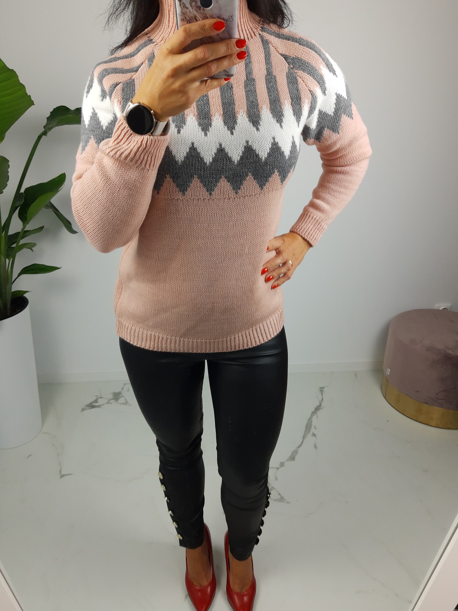 Kobieta w różowym swetrze z geometrycznym wzorem, czarnych skórzanych spodniach z ozdobnymi guzikami i czerwonych szpilkach, robiąca sobie zdjęcie w lustrze, widoczny zegarek na ręce.