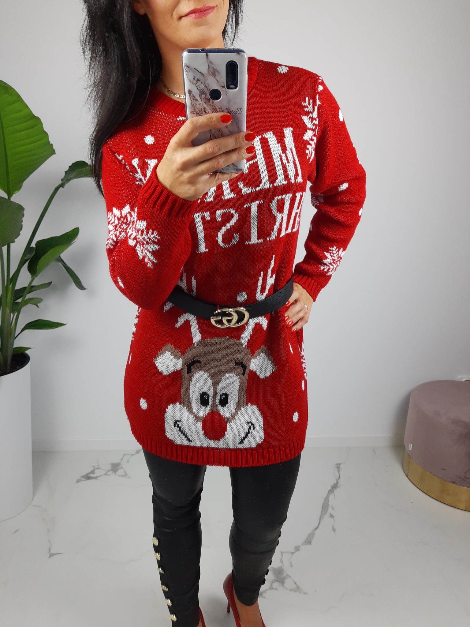 Kobieta robi selfie w czerwonym świątecznym swetrze z reniferem, czarnych skórzanych spodniach z guzikami i czerwonych szpilkach, stojąc na marmurowej podłodze obok rośliny doniczkowej.