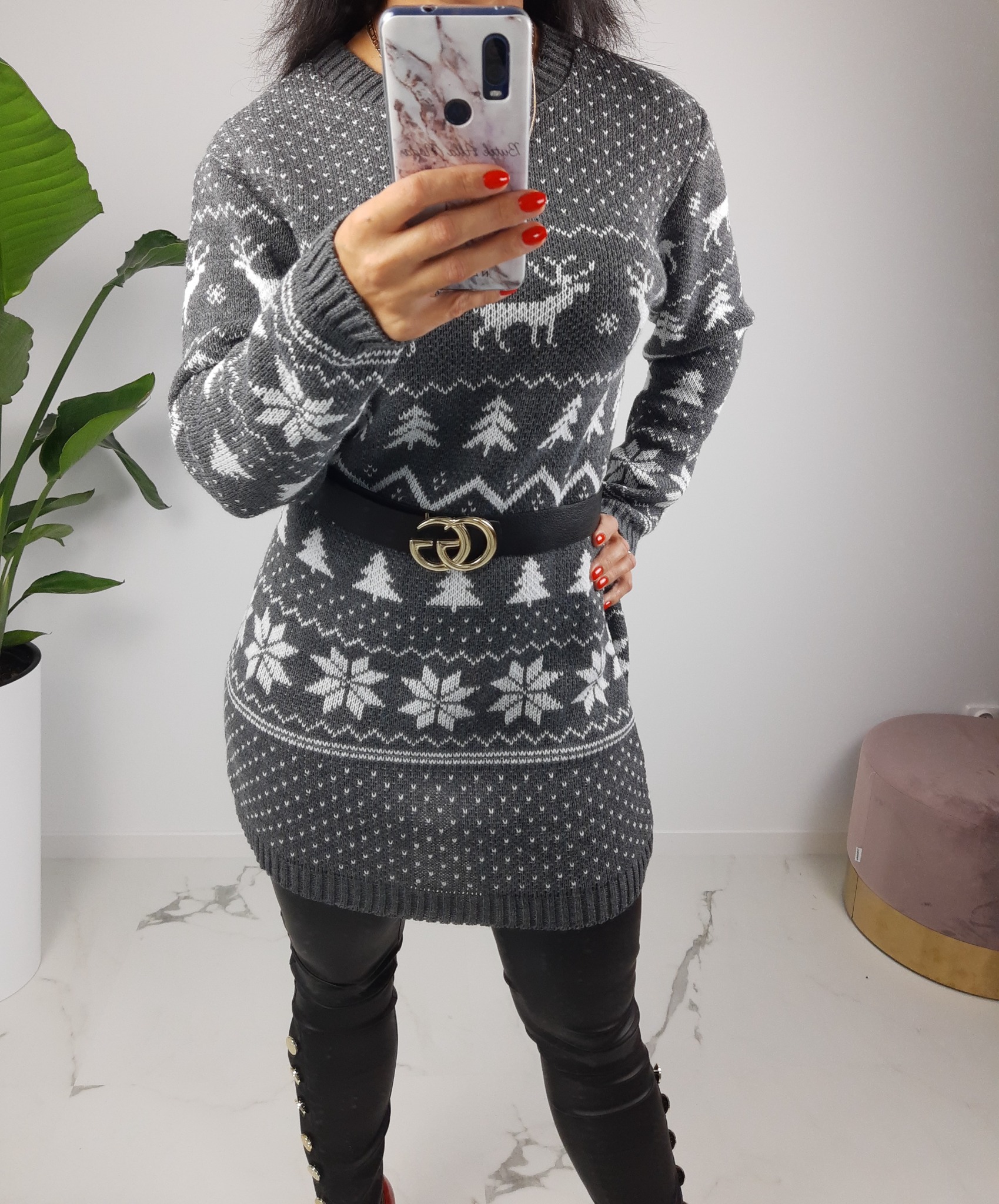 Kobieta robi selfie w szarej dzianinowej tunice z wzorem reniferów i choinek, przewiązana czarnym paskiem z złotą klamrą Gucci, w skórzanych spodniach z guzikami po bokach.