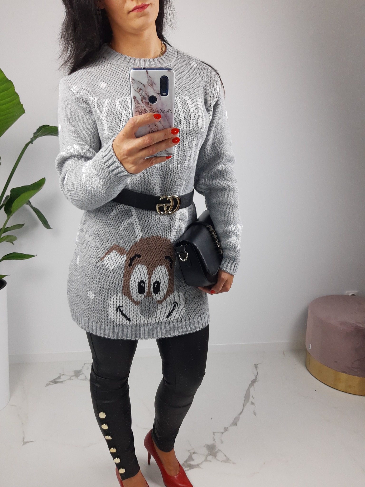 Kobieta robi selfie w szarym swetrze z reniferem, czarnych legginsach z guzikami i czerwonych szpilkach, trzymając czarną torebkę.