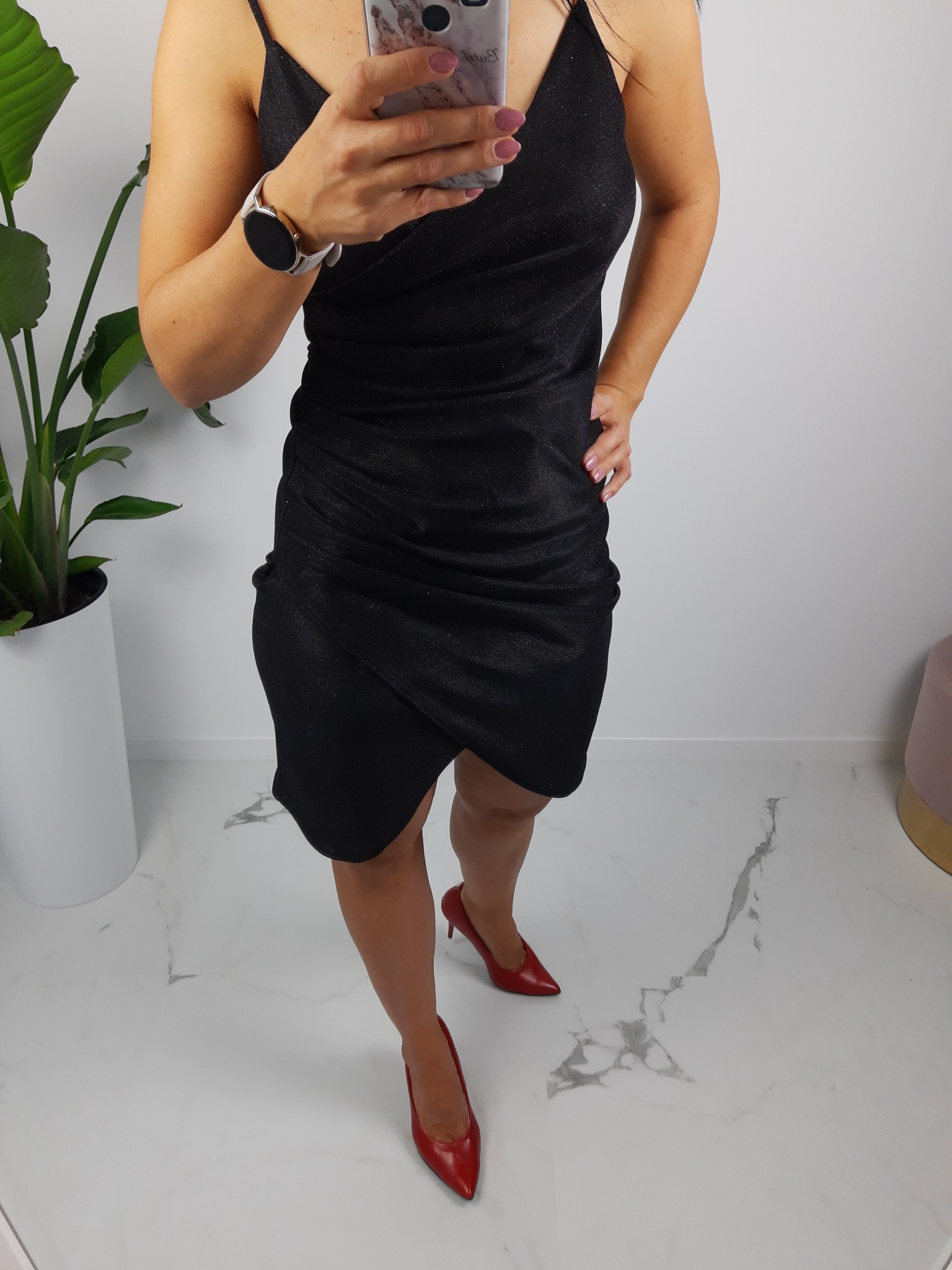 Kobieta w czarnej, błyszczącej sukience na ramiączkach, pozująca z telefonem i czerwonych szpilkach na marmurowej podłodze, obok rośliny doniczkowej.