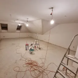 Poddasze w trakcie prac wykończeniowych: surowe ściany, dwa okna dachowe, prowizoryczne oświetlenie na kablach, dwie drabiny malarskie, wiaderka z farbą i zwój kabli na podłodze.