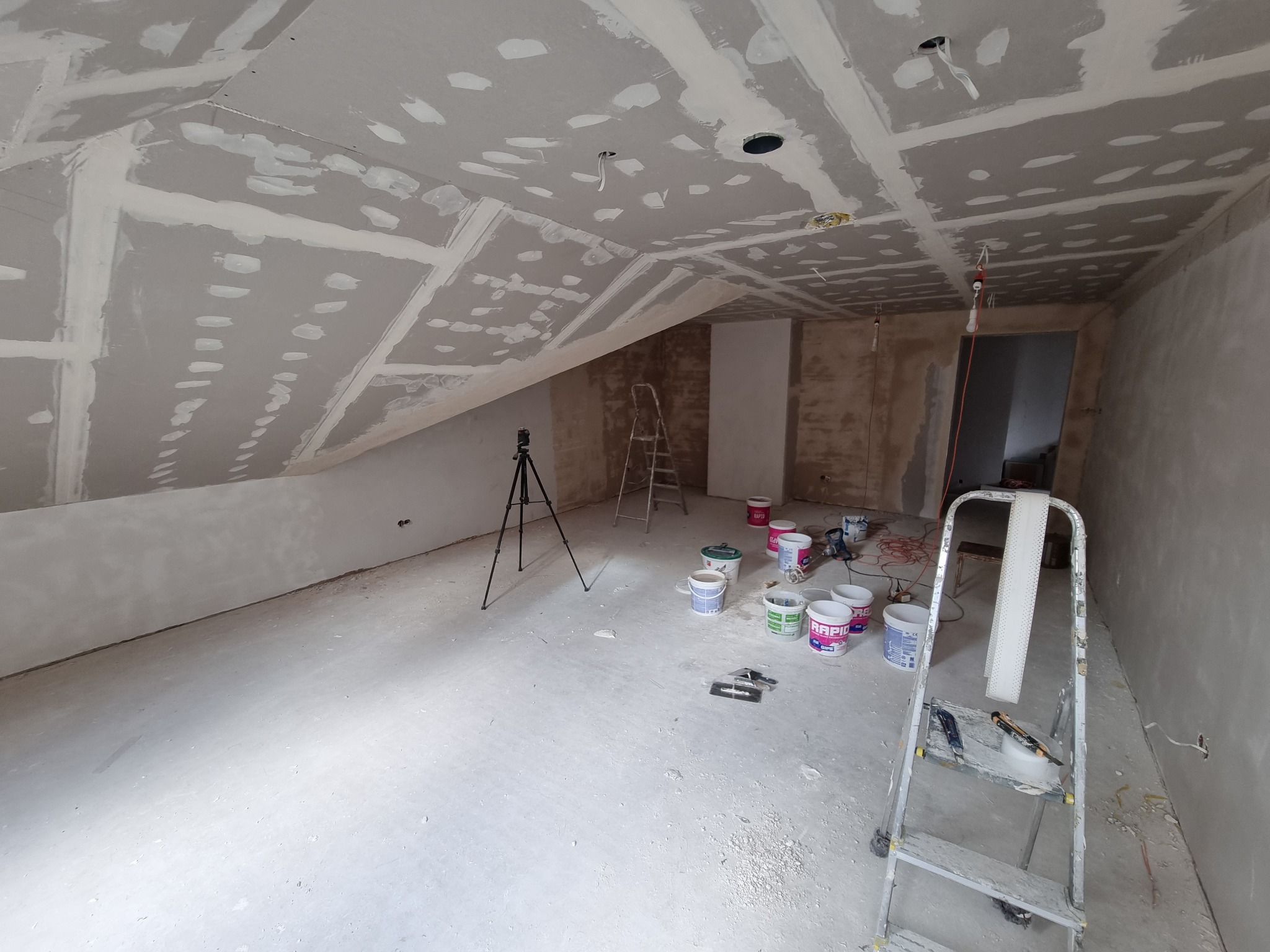 Poddasze w trakcie szpachlowania płyt gipsowo-kartonowych, widoczne łączenia płyt wypełnione gładzią, drabina, puszki z farbą i narzędzia malarskie.