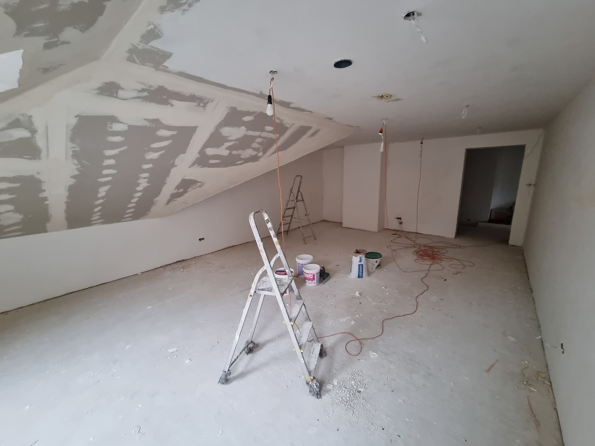 Pokój na poddaszu w trakcie szpachlowania i przygotowania do malowania, widoczna drabina, wiadra z farbą, sufit ze śladami szpachli i wystającymi kablami. Na podłodze widoczne resztki gipsu.