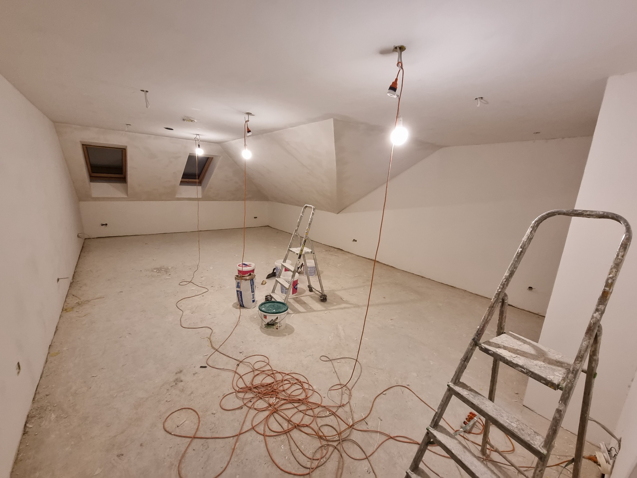 Poddasze w trakcie prac wykończeniowych: surowe ściany, dwa okna dachowe, prowizoryczne oświetlenie na kablach, dwie drabiny malarskie, wiaderka z farbą i zwój kabli na podłodze.