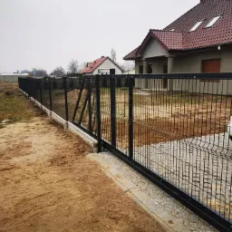 Czarny panelowy system ogrodzeniowy z przesuwną bramą, zamontowany na betonowej podmurówce, widok na dom i zaparkowany samochód w pochmurny dzień.