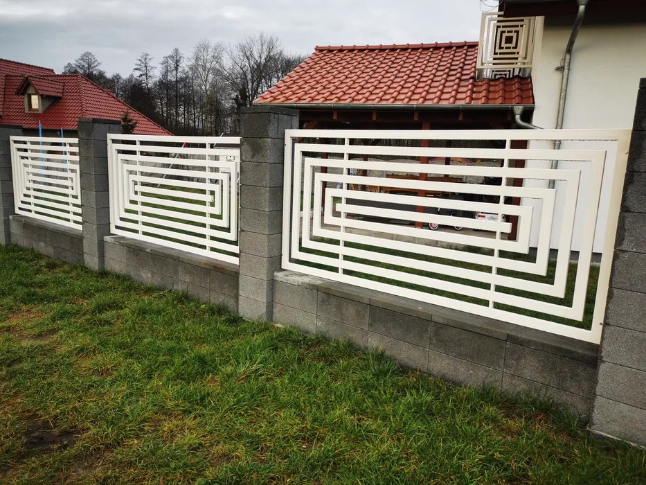 Nowoczesne, białe ogrodzenie metalowe z geometrycznym wzorem na betonowej podmurówce, widok z poziomu trawnika na tle zabudowań.