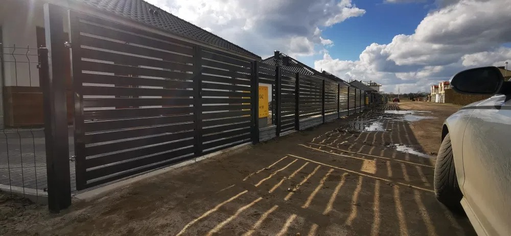 Nowoczesne, poziome, czarne ogrodzenie metalowe z Sulechowa, widoczne z perspektywy ulicy z błotem i odbiciami światła, częściowo zasłonięte przez samochód.