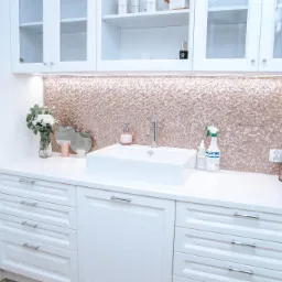 Biała szafka łazienkowa z umywalką nablatową, baterią, mozaiką w odcieniach różowego złota, kwiatami w wazonie i kosmetykami, oświetlona listwą LED pod szafkami wiszącymi.