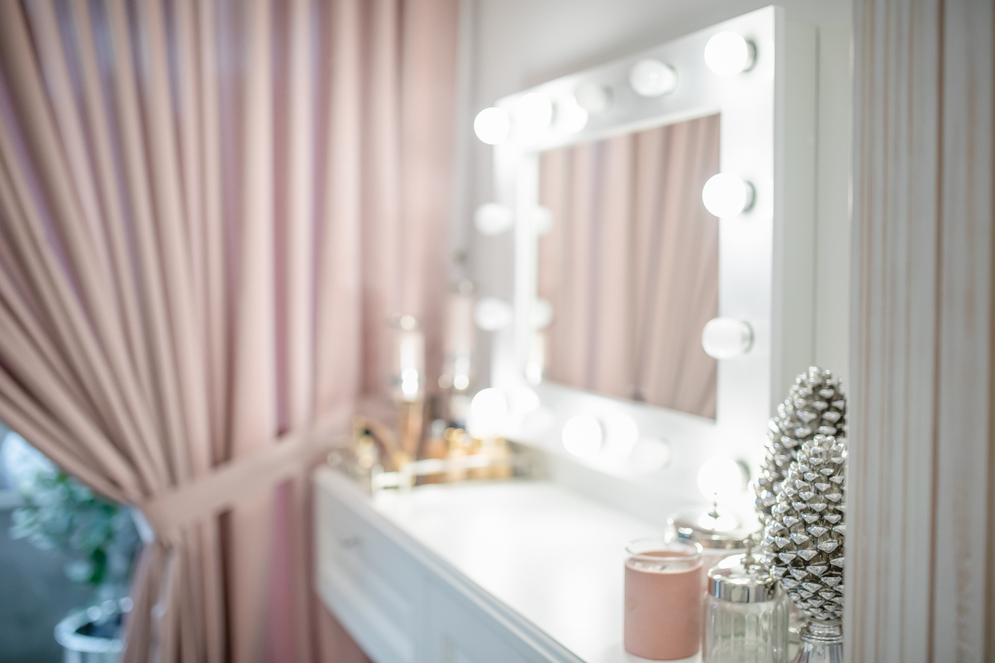 Toaletka z białym lustrem otoczonym żarówkami, dekorowana srebrnymi szyszkami i różową zasłoną w tle.
